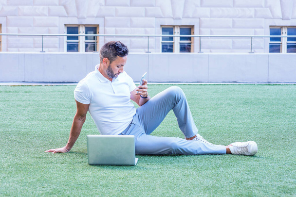 Young European Man with beard, little gray hair, wearing white short sleeve shirt, gray pants, white sneakers, sitting on green lawn in New York City, working on laptop computer, texting on cell phon - Photo, Image