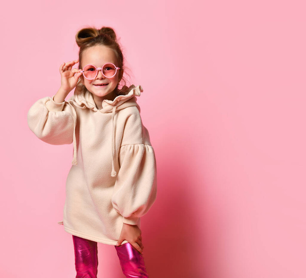 Little blonde in glasses, a stylish sweatshirt and leggings. Looking at you intently or with a smile, posing on a pink studio background. - Фото, зображення