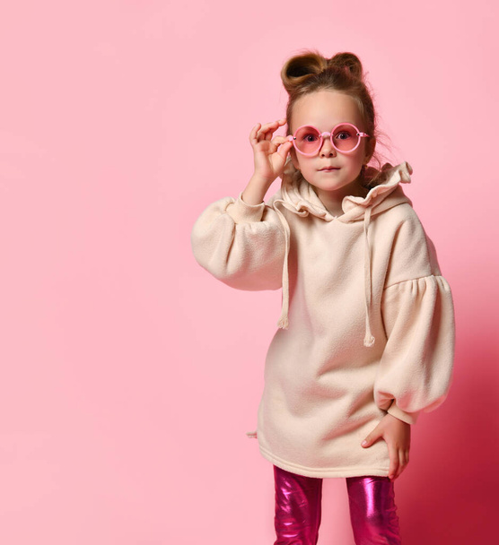 Little blonde girl in glasses, stylish hoodie and leggings. Looking at you intently or scared, posing on pink studio background. Close up, copy space - Φωτογραφία, εικόνα