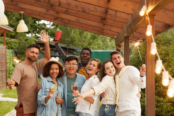 Grupo multiétnico de jóvenes despreocupados sosteniendo bengalas y sonriendo a la cámara mientras disfrutan de la fiesta de verano en la terraza al aire libre, espacio para copiar - Foto, Imagen