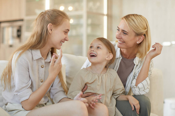 Nettes warmgetöntes Porträt einer unbeschwerten jungen Mutter, die mit zwei Töchtern spricht und fröhlich lächelt, während sie die gemeinsame Zeit zu Hause genießt - Foto, Bild