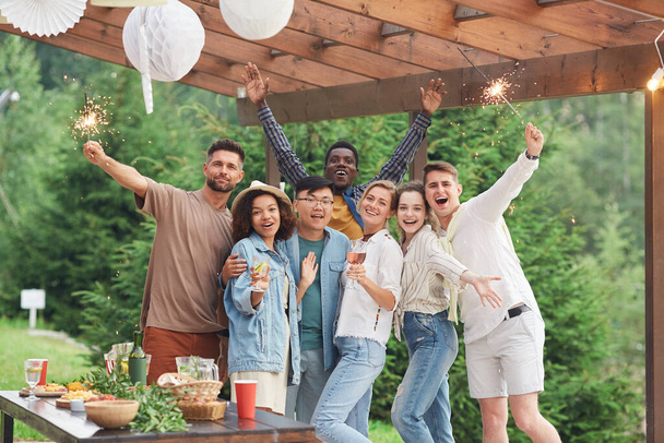Grupo multi-étnico de amigos felizes segurando sparklers e olhando para a câmera enquanto desfruta de festa de verão ao ar livre, espaço de cópia - Foto, Imagem