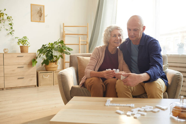Caliente retrato tonificado de feliz pareja de ancianos sosteniendo piezas de dominó mientras juega juegos de mesa en casa, espacio de copia - Foto, imagen