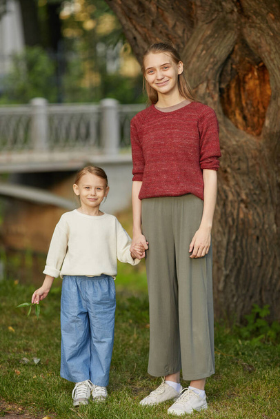 Vertical full length portrait of two sisters holding hands while standing in park outdoors and looking at camera - Foto, afbeelding