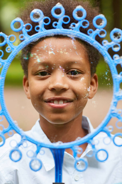 Ritratto verticale ravvicinato di un ragazzo afro-americano sorridente che guarda la fotocamera attraverso una grande bacchetta di bolla mentre gioca all'aperto nel parco - Foto, immagini