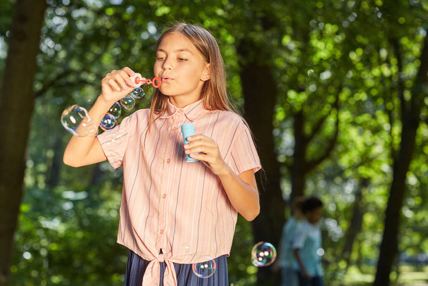 Waist up portrait of teenage girl blowing bubbles while standing in green park outdoors and lit by sunlight, copy space - Фото, изображение