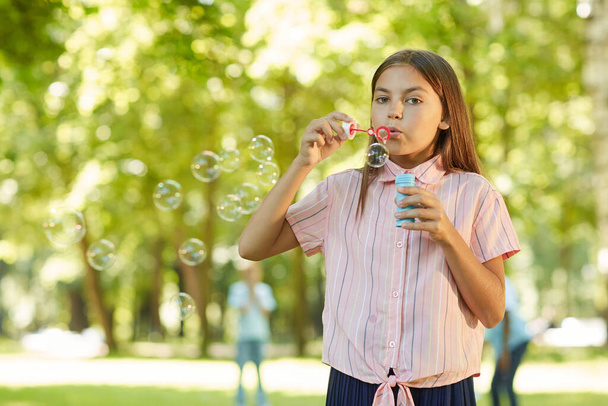 Derék fel portré tinédzser lány fúj buborékok és a kamerába, miközben áll a zöld parkban a szabadban, másolja a helyet - Fotó, kép