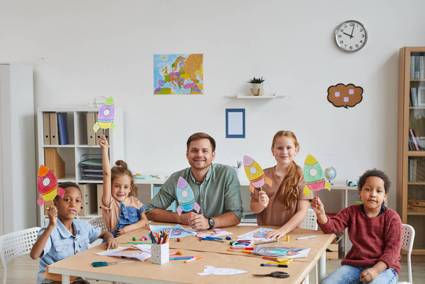 Retrato de un profesor sonriente con un grupo multiétnico de niños mostrando imágenes de cohetes espaciales mientras disfruta de clases de arte y artesanía en preescolar o centro de desarrollo, espacio para copiar - Foto, Imagen