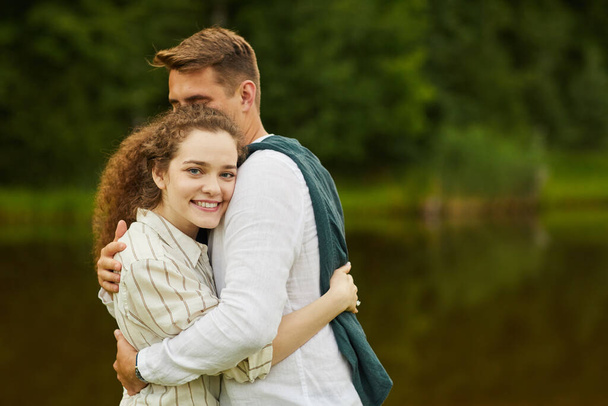Waist up portrait of loving young couple embracing and looking at camera while standing by lake outdoors in Summer, copy space - Photo, Image
