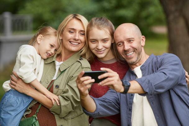 taille omhoog portret van moderne gelukkige familie met twee dochters selfie buiten via smartphone onder het genot van wandeling in groen park - Foto, afbeelding