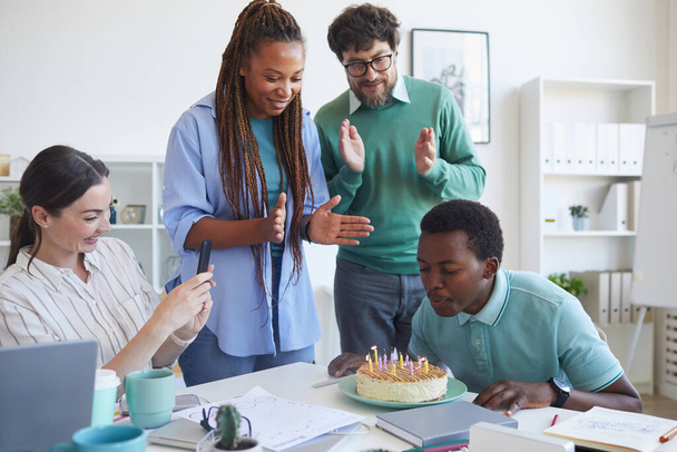 Мульти-этническая группа людей, празднующих День рождения в офисе, сосредоточиться на молодой афроамериканец задувая свечи на торте с коллегами аплодируя, копировать пространство - Фото, изображение