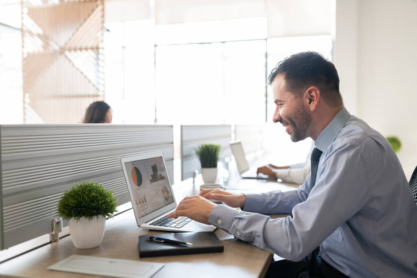 Usmívající se podnikatel pracující na notebooku, zatímco sedí za stolem v kanceláři - Fotografie, Obrázek