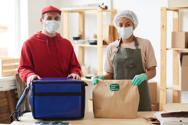 Талия портрет двух рабочих в масках и смотреть в камеру во время упаковки заказов в бесконтактной службы доставки продуктов питания, копировать пространство - Фото, изображение