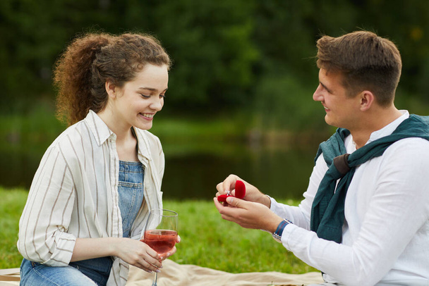Vista lateral en la caja de anillo de apertura de hombre joven mientras que propone a la novia durante la cita romántica al aire libre - Foto, imagen