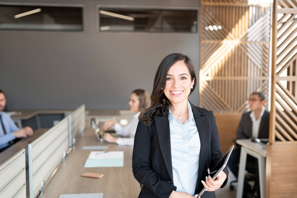 Портрет жінки-юриста, що тримає цифровий планшет, що стоїть в офісі з колегами, які працюють у фоновому режимі
 - Фото, зображення