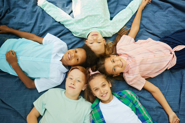 Vue de dessus du groupe multi-ethnique d'enfants allongés en cercle sur une couverture à l'extérieur et regardant la caméra - Photo, image