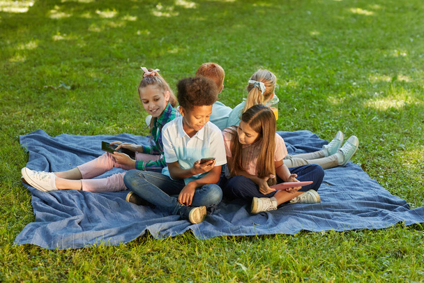 Άποψη υψηλής γωνίας σε πολυεθνική ομάδα παιδιών που χρησιμοποιούν gadgets ενώ κάθονται σε πράσινο γρασίδι σε εξωτερικούς χώρους πάρκο, αντιγραφή χώρου - Φωτογραφία, εικόνα