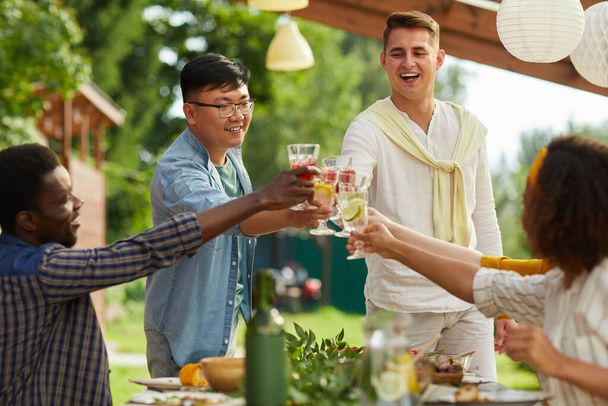 Multietnická skupina přátel si užívá večeři venku na letním večírku, zaměřují se na dva muže cinkající sklenicemi při stání u stolu, kopírují prostor - Fotografie, Obrázek