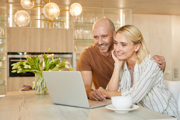 Warm-toned portrait of happy adult couple looking at laptop screen and smiling while talking by video chat with family, copy space - Fotografie, Obrázek