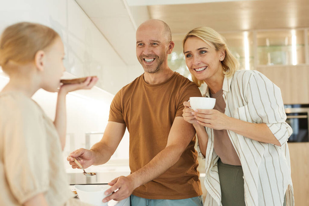 Cintura cálida hacia arriba retrato de padres felices mirando linda niña que alivia el desayuno saludable en el interior de la cocina moderna - Foto, imagen