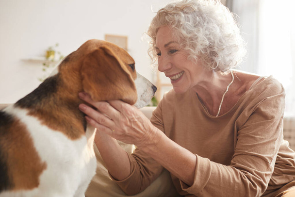 Seitenansicht Porträt einer weißhaarigen Seniorin, die mit Hund spielt und lächelt, während sie auf der Couch im gemütlichen Hausinneren sitzt, Kopierraum - Foto, Bild