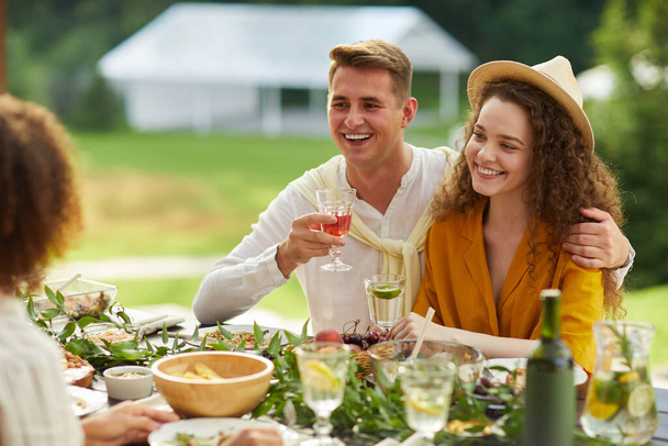 Portré fiatal pár ölelkezik, miközben ül asztal kezében italok és élvezi a vacsorát barátaival a szabadban a nyári party, másolás tér - Fotó, kép