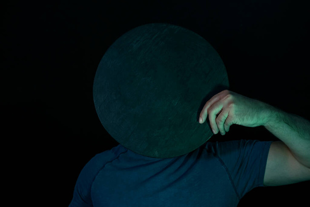 Young guy covered his face with a black round disk in neon light on a black. Minimalism retro style concept. - Photo, Image
