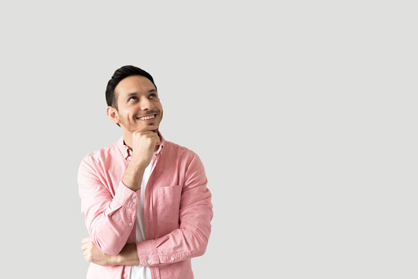 Латиноамериканец в повседневной одежде смотрит вверх в сторону копировального пространства и улыбается в студии - Фото, изображение