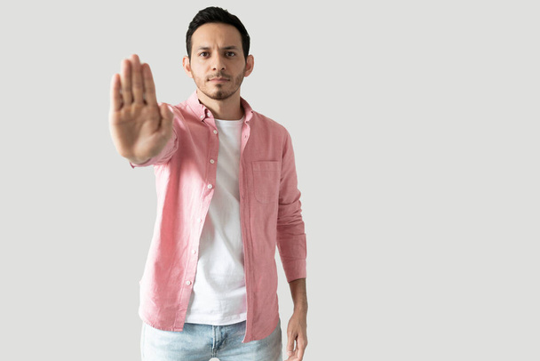 Hombre de aspecto serio sosteniendo su mano abierta y haciendo un gesto de stop en un estudio sobre un fondo gris - Foto, imagen