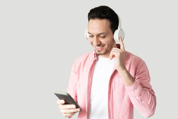 Νεαρός άνδρας ντυμένος casual και ακούγοντας μουσική με ασύρματα ακουστικά και ένα smartphone σε στούντιο - Φωτογραφία, εικόνα