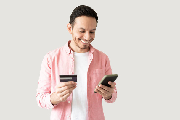 Aantrekkelijke Latijnse jongeman met een creditcard en een smartphone om wat spullen online te kopen - Foto, afbeelding