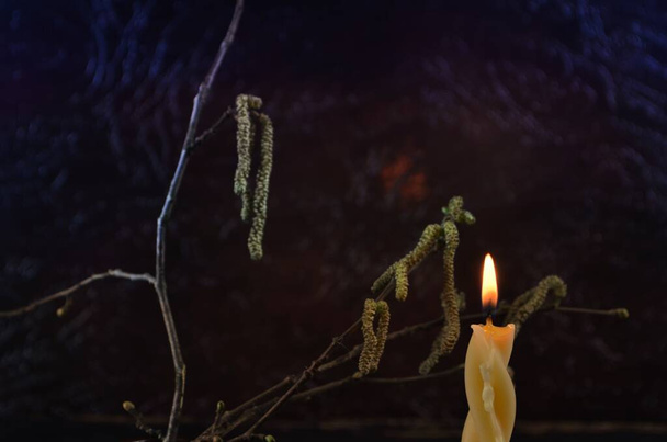 Candela di cera ardente nel portacandele in argilla e ramo di nocciola in fiore sullo sfondo, sfondo scuro - Foto, immagini