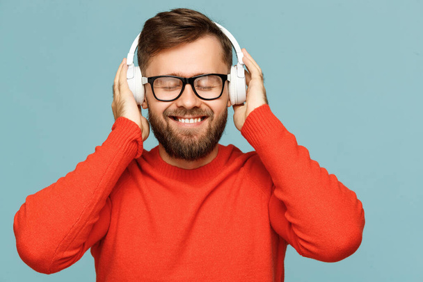 Vrolijke man met een baard in een bril glimlachend en luisterend naar goede muziek in een koptelefoon met gesloten ogen tegen een blauwe achtergrond - Foto, afbeelding