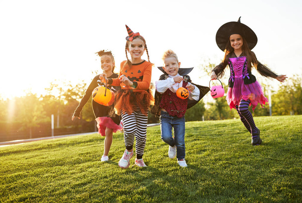 Grupo de crianças animadas em fantasias assustadoras sorrindo e correndo no gramado durante a celebração de Halloween à noite, a par - Foto, Imagem