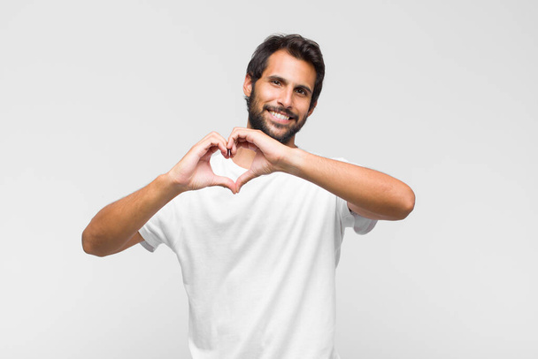 young latin handsome man smiling and feeling happy, cute, romantic and in love, making heart shape with both hands - Foto, Imagem
