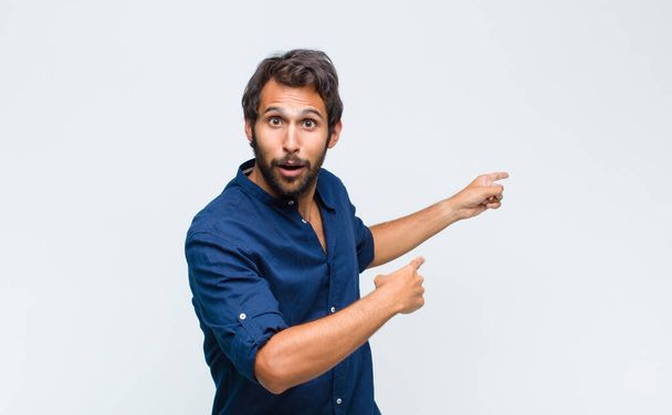 young latin handsome man feeling shocked and surprised, pointing to copy space on the side with amazed, open-mouthed look - Фото, зображення
