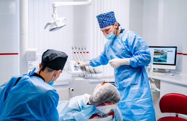 Male dentist is working in modern stomatology clinic. Stomatological instrument in the dentist clinic. Patient in dentist chair. Assistant in dentistry. - Fotó, kép