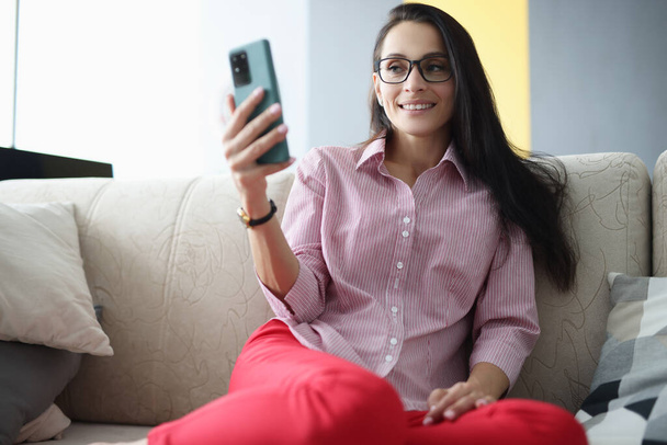 Donna con gli occhiali siede sul divano sorridente e guarda lo schermo dello smartphone - Foto, immagini
