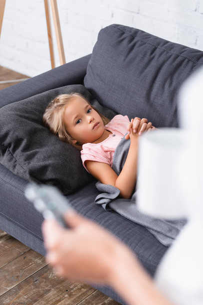 Concentration sélective de l'enfant malade couché sur le canapé près de la mère avec tasse et pilules  - Photo, image
