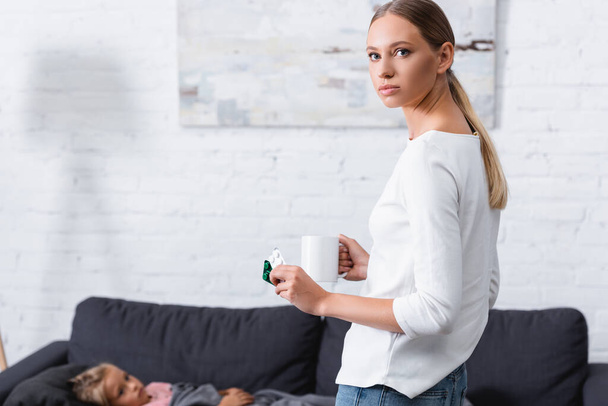 Selective focus of young woman with mug and pills looking at camera near sick child on couch at home  - Fotografie, Obrázek
