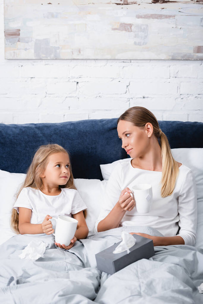 Enfoque selectivo de la madre y el niño mirándose mientras sostienen tazas cerca de servilletas en la cama  - Foto, Imagen