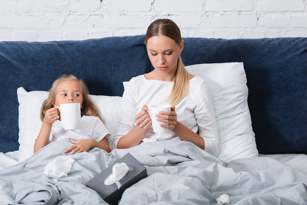Selective focus of woman holding cup near sick child and clumped napkins on bed  - Foto, imagen