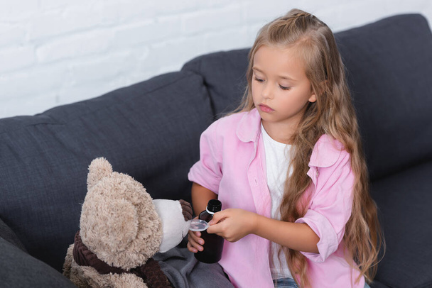 Girl holding spoon and bottle of syrup near soft toy on couch at home  - Фото, зображення