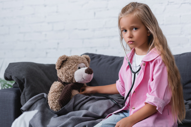 Girl in stethoscope looking at camera near teddy bear on couch  - Zdjęcie, obraz