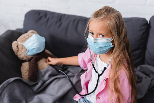 Selective focus of girl in medical mask holding stethoscope near teddy bear  - Foto, afbeelding