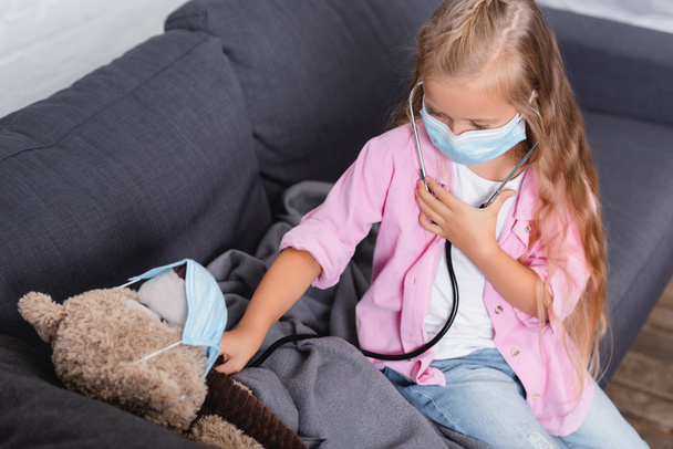 Selectieve focus van meisje in medisch masker houden stethoscoop in de buurt van zacht speelgoed thuis  - Foto, afbeelding