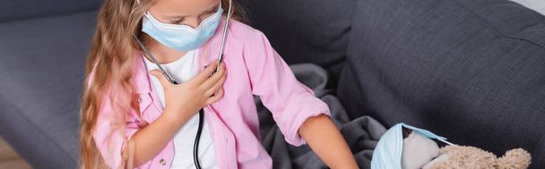 Immagine orizzontale di bambino in maschera medica in stetoscopio seduto vicino orsacchiotto sul divano  - Foto, immagini