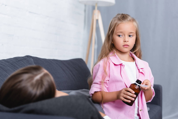 Selective focus of child holding spoon and syrup near diseased mother on couch  - Фото, изображение