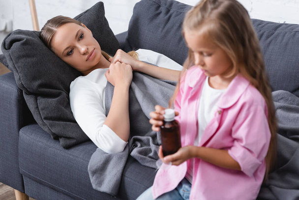 Επιλεκτική εστίαση της άρρωστης γυναίκας κοιτάζοντας την κόρη με ένα μπουκάλι σιρόπι στο σπίτι  - Φωτογραφία, εικόνα