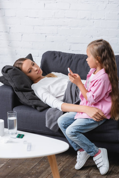 Selective focus of sick woman embracing kid with thermometer near pills and water on coffee table  - Photo, Image